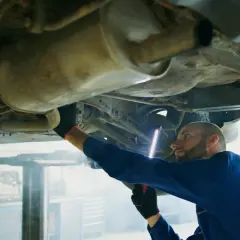 Mechanik w niebieskim kombinezonie bada podwozie auta, używając latarki. To typowa scena z warsztatu, gdzie każdy szczegół, jak ten z p0420, jest ważny.