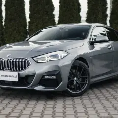 Szary BMW 218i z czarnymi felgami stoi przed żywopłotem.