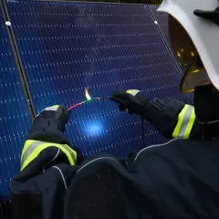 Strażak w białym hełmie i rękawicach tworzy łuk elektryczny, zbliżając dwa przewody do paneli słonecznych.