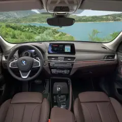 Wnętrze BMW X1 z brązową tapicerką i ekranem nawigacji. Widok na jezioro i góry. Sprawdź aktualną bmw x1 cena.