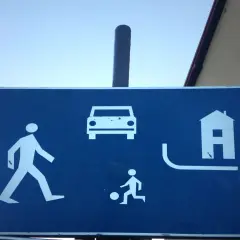 Niebieski znak informuje o strefie zamieszkania: piesi, samochody, dzieci grające w piłkę i domy.