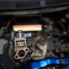 Zardzewiały zawór EGR, który może być przyczyną problemów z silnikiem. Mechanik pokazuje go na tle komory silnika.