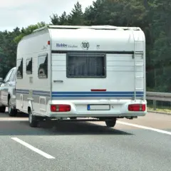 Biała przyczepa kempingowa Hobby z niebieskim pasem jedzie autostradą. Kierowca z prawem jazdy kategorii B+E wiezie rodzinę na wakacje.