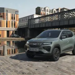 Dacia Spring cena: zielony elektryczny samochód miejski stoi na brukowanej ulicy nad kanałem, w tle nowoczesne budynki i most.