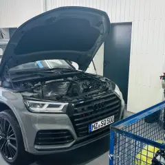 Szary Audi Q7 3.0 TDI z otwartą maską i bagażnikiem w warsztacie. Obok stoi wózek z narzędziami i silnik.