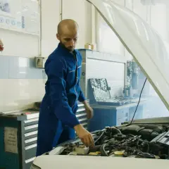 Mechanik w niebieskim kombinezonie sprawdza silnik samochodu, z którego wydobywa się dym. Obok stoi skrzynka z narzędziami i urządzenie przypominające parownik.