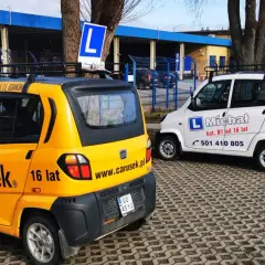 Dwa samochody szkoleniowe, żółty i biały, z literą "L" na dachu. Na żółtym aucie napis "Carusek 16 lat", sugerujący możliwość uzyskania prawo jazdy od 16 lat.