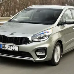 Srebrny Kia Carens 4 w ruchu drogowym, z widocznym grillem i reflektorami.