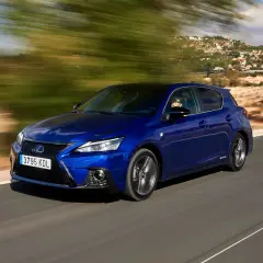 Błękitny Lexus CT 200h pędzi po krętej drodze, mijając zielone drzewa i domy na wzgórzu.