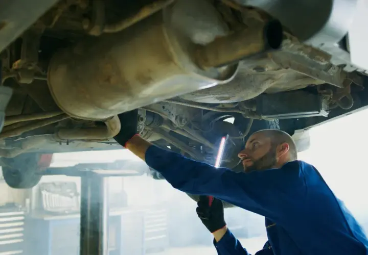 Mechanik w niebieskim kombinezonie bada podwozie auta, używając latarki. To typowa scena z warsztatu, gdzie każdy szczegół, jak ten z p0420, jest ważny.