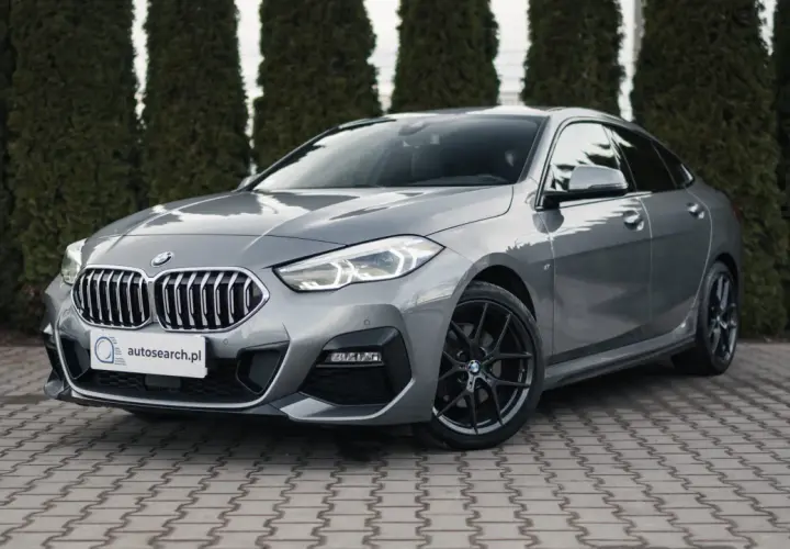 Szary BMW 218i z czarnymi felgami stoi przed żywopłotem.