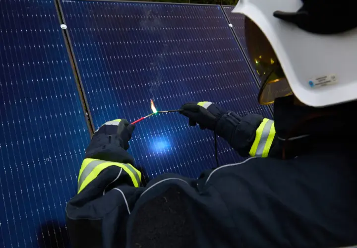 Strażak w białym hełmie i rękawicach tworzy łuk elektryczny, zbliżając dwa przewody do paneli słonecznych.