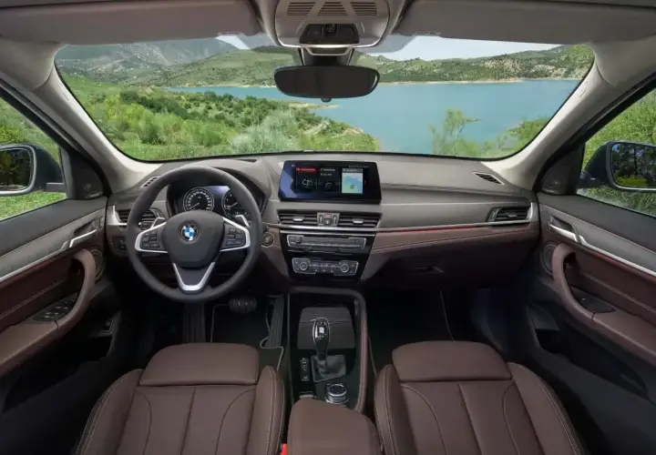 Wnętrze BMW X1 z brązową tapicerką i ekranem nawigacji. Widok na jezioro i góry. Sprawdź aktualną bmw x1 cena.