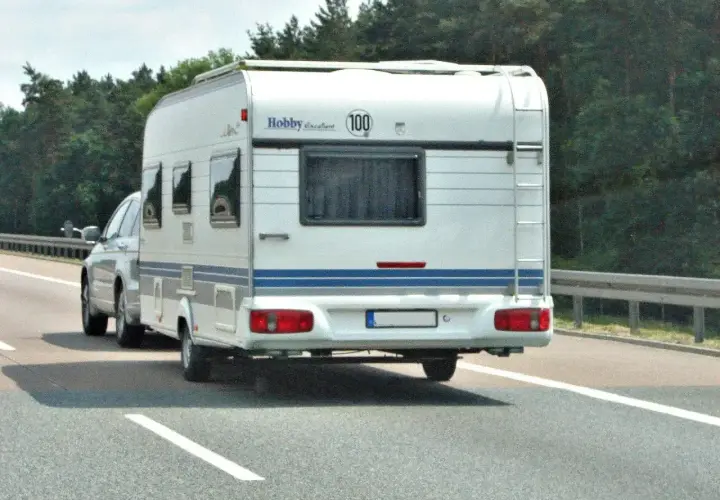 Biała przyczepa kempingowa Hobby z niebieskim pasem jedzie autostradą. Kierowca z prawem jazdy kategorii B+E wiezie rodzinę na wakacje.