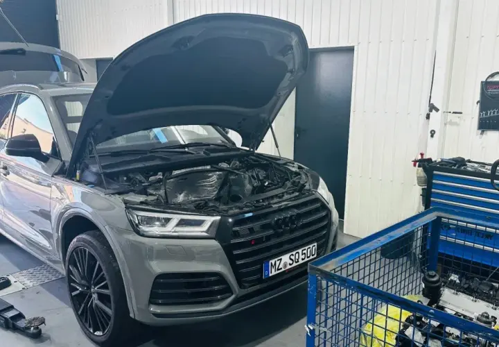Szary Audi Q7 3.0 TDI z otwartą maską i bagażnikiem w warsztacie. Obok stoi wózek z narzędziami i silnik.