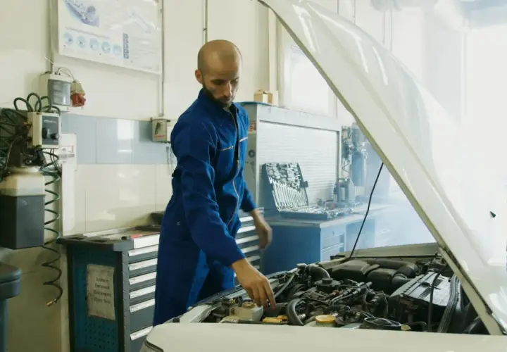 Mechanik w niebieskim kombinezonie sprawdza silnik samochodu, z którego wydobywa się dym. Obok stoi skrzynka z narzędziami i urządzenie przypominające parownik.