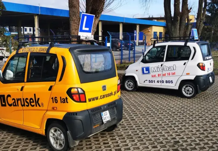 Dwa samochody szkoleniowe, żółty i biały, z literą "L" na dachu. Na żółtym aucie napis "Carusek 16 lat", sugerujący możliwość uzyskania prawo jazdy od 16 lat.