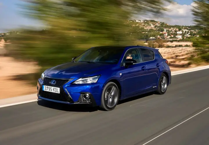 Błękitny Lexus CT 200h pędzi po krętej drodze, mijając zielone drzewa i domy na wzgórzu.