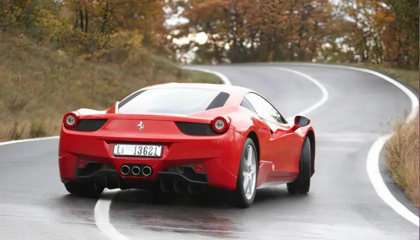 Czerwone Ferrari 458 na mokrej, krętej drodze wśród jesiennych drzew.
