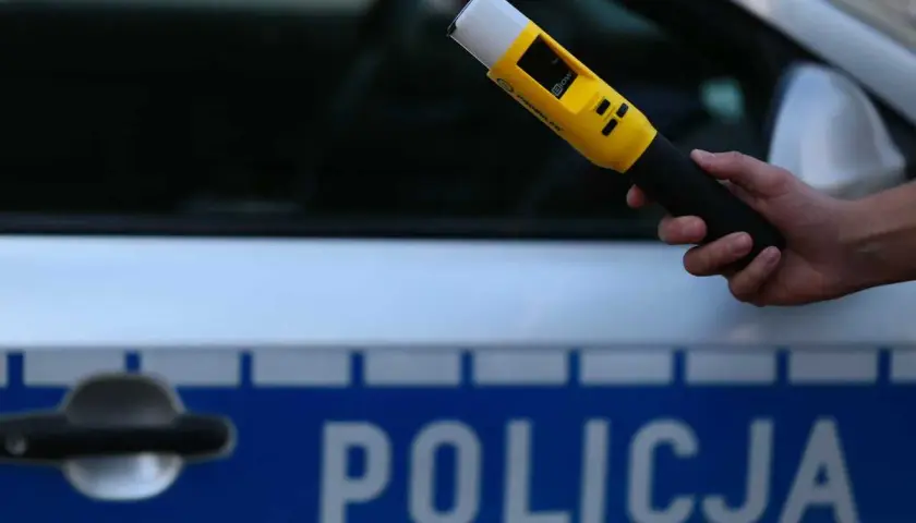Dłoń trzymająca żółty alkomat na tle radiowozu policyjnego z napisem "POLICJA".