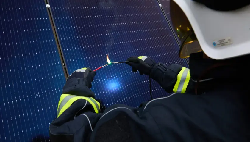 Strażak w białym hełmie i rękawicach tworzy łuk elektryczny, zbliżając dwa przewody do paneli słonecznych.