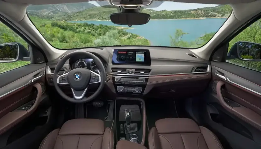 Wnętrze BMW X1 z brązową tapicerką i ekranem nawigacji. Widok na jezioro i góry. Sprawdź aktualną bmw x1 cena.