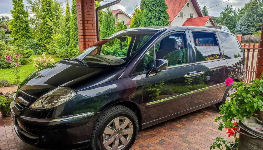 Czarny Citroen C8 zaparkowany pod zadaszeniem, otoczony zielenią i domami z czerwonymi dachami.