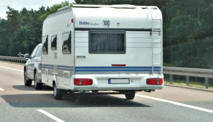 Biała przyczepa kempingowa Hobby z niebieskim pasem jedzie autostradą. Kierowca z prawem jazdy kategorii B+E wiezie rodzinę na wakacje.