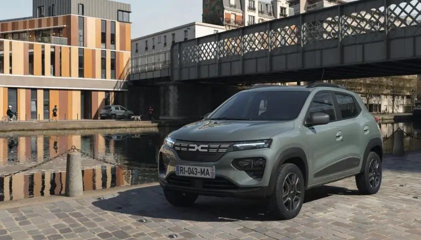 Dacia Spring cena: zielony elektryczny samochód miejski stoi na brukowanej ulicy nad kanałem, w tle nowoczesne budynki i most.