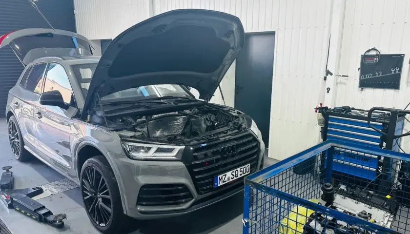 Szary Audi Q7 3.0 TDI z otwartą maską i bagażnikiem w warsztacie. Obok stoi wózek z narzędziami i silnik.