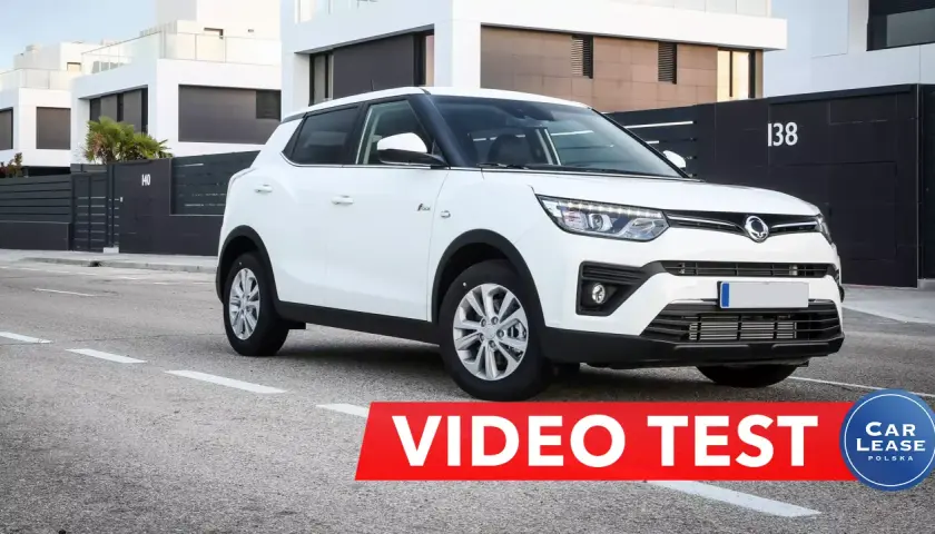 Biały tivoli samochód na tle nowoczesnych domów. Czerwony pas z napisem "VIDEO TEST" i logo "CAR LEASE POLSKA".