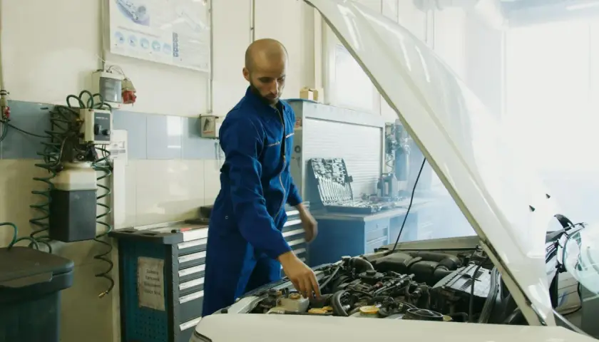 Mechanik w niebieskim kombinezonie sprawdza silnik samochodu, z którego wydobywa się dym. Obok stoi skrzynka z narzędziami i urządzenie przypominające parownik.