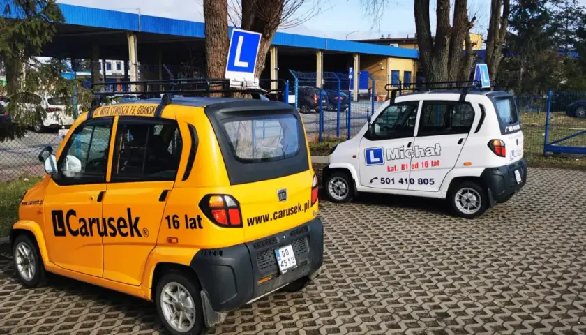 Dwa samochody szkoleniowe, żółty i biały, z literą "L" na dachu. Na żółtym aucie napis "Carusek 16 lat", sugerujący możliwość uzyskania prawo jazdy od 16 lat.
