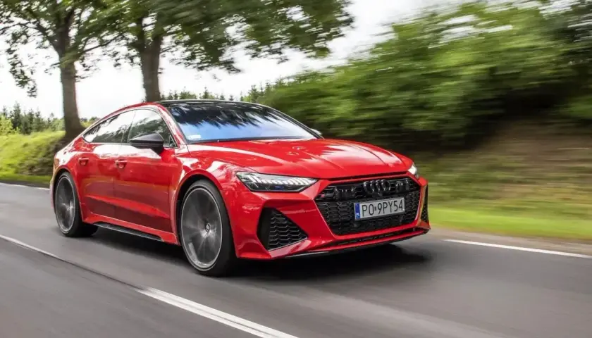 Czerwony Audi RS7 Sportback pędzi po drodze, otoczony zielenią drzew.