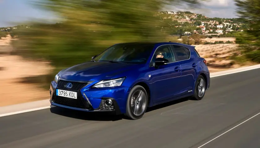 Błękitny Lexus CT 200h pędzi po krętej drodze, mijając zielone drzewa i domy na wzgórzu.