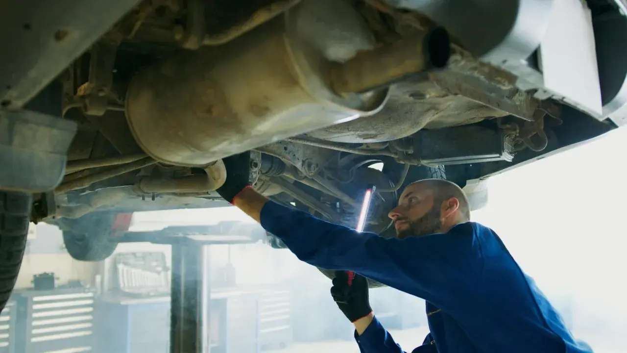 Mechanik w niebieskim kombinezonie bada podwozie auta, używając latarki. To typowa scena z warsztatu, gdzie każdy szczegół, jak ten z p0420, jest ważny.