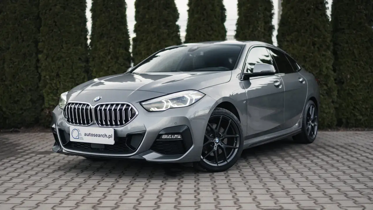 Szary BMW 218i z czarnymi felgami stoi przed żywopłotem.