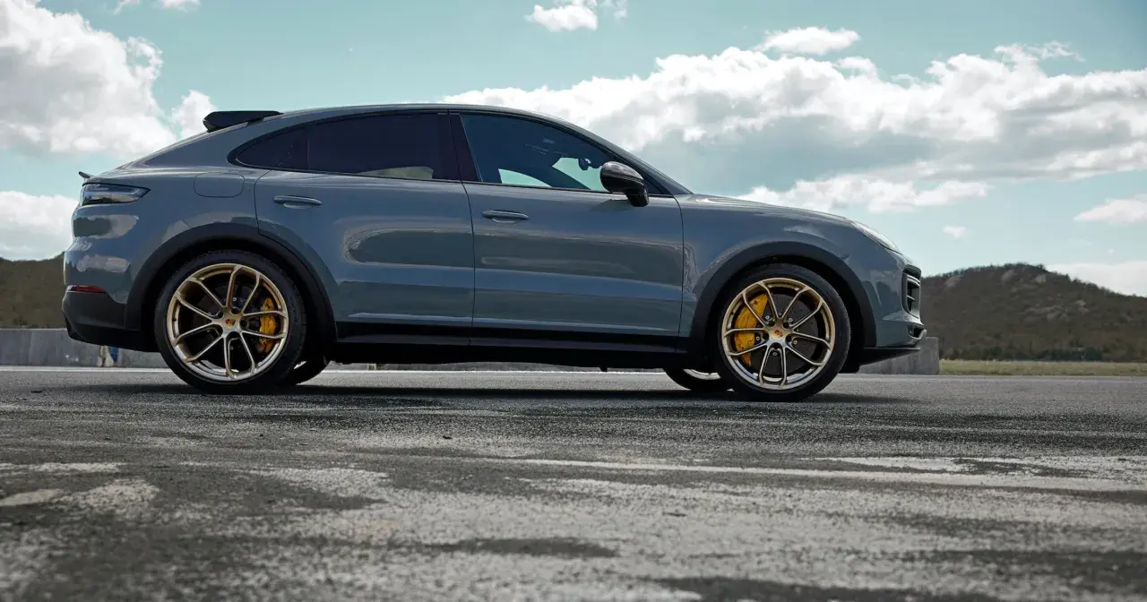 Szary Porsche Cayenne Turbo GT z żółtymi zaciskami hamulcowymi na złotych felgach, zaparkowany na pustej drodze z górzystym krajobrazem w tle.