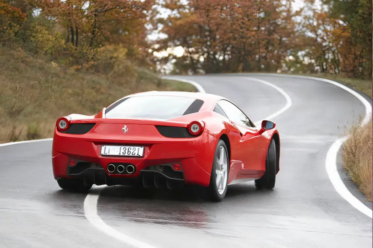 Czerwone Ferrari 458 na mokrej, krętej drodze wśród jesiennych drzew.