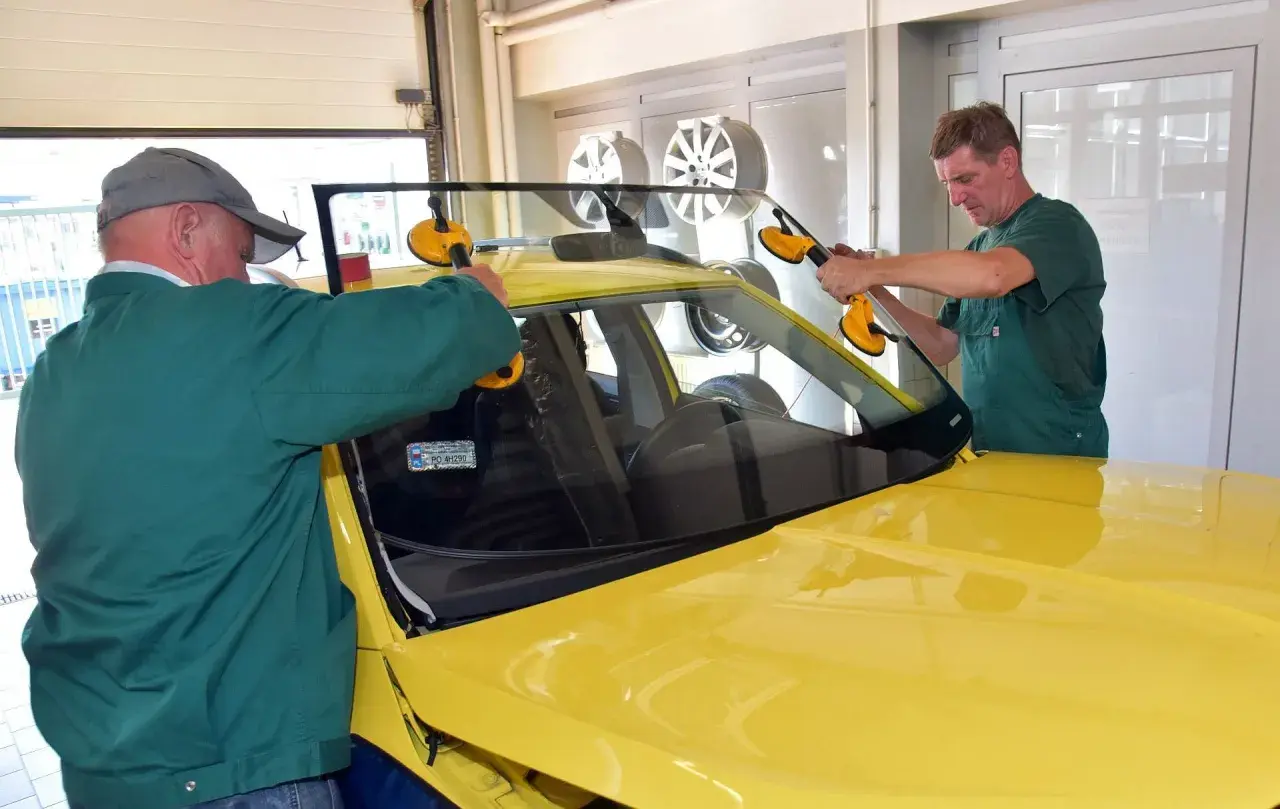 Dwóch mechaników montuje przednią szybę w żółtym samochodzie. Sprawdź autoglass cennik usług.