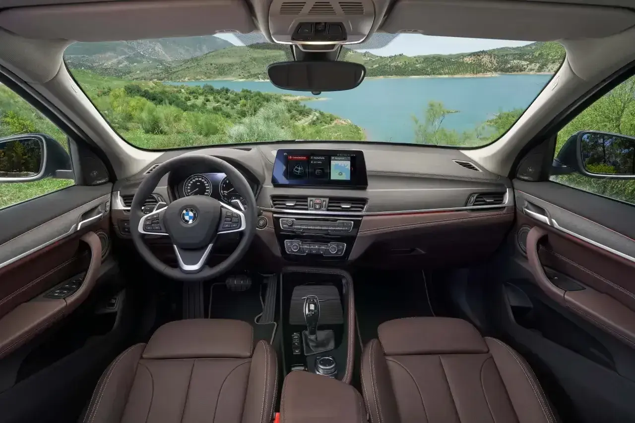Wnętrze BMW X1 z brązową tapicerką i ekranem nawigacji. Widok na jezioro i góry. Sprawdź aktualną bmw x1 cena.