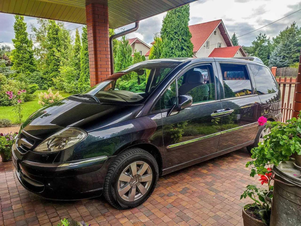 Czarny Citroen C8 zaparkowany pod zadaszeniem, otoczony zielenią i domami z czerwonymi dachami.