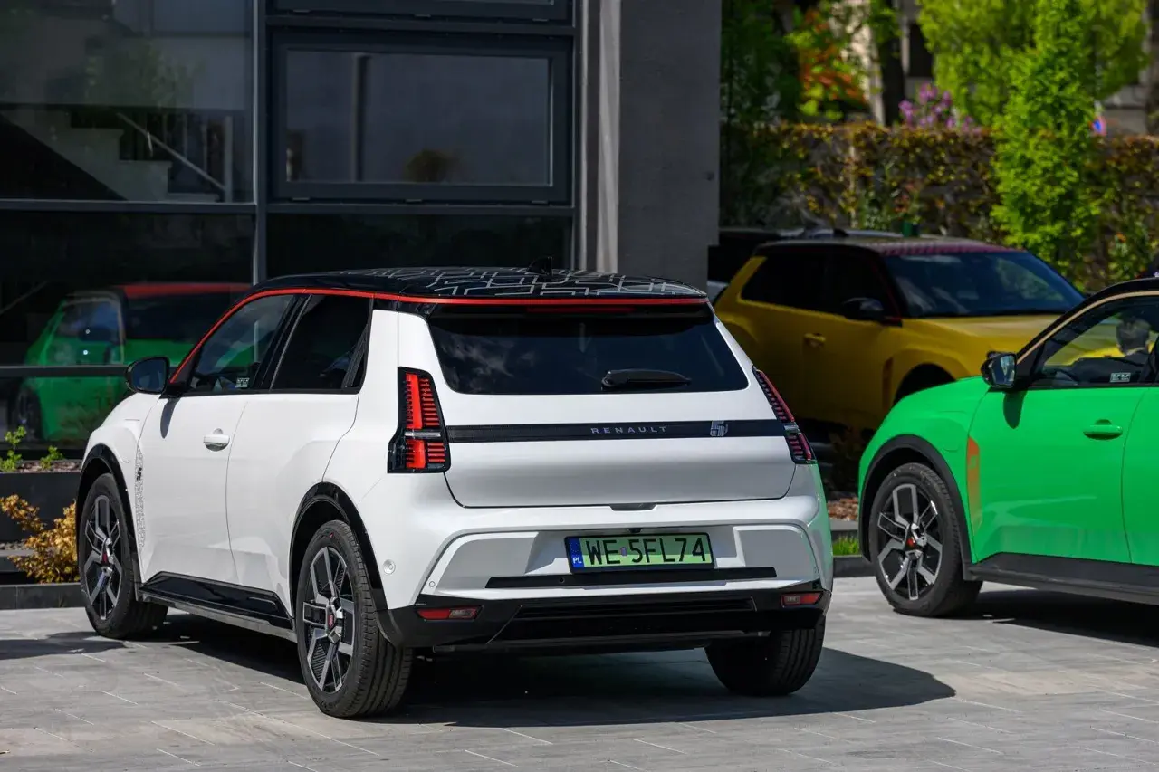 Biały Renault 5 e-tech z czarnym dachem i czerwonymi akcentami stoi obok zielonego i żółtego samochodu.