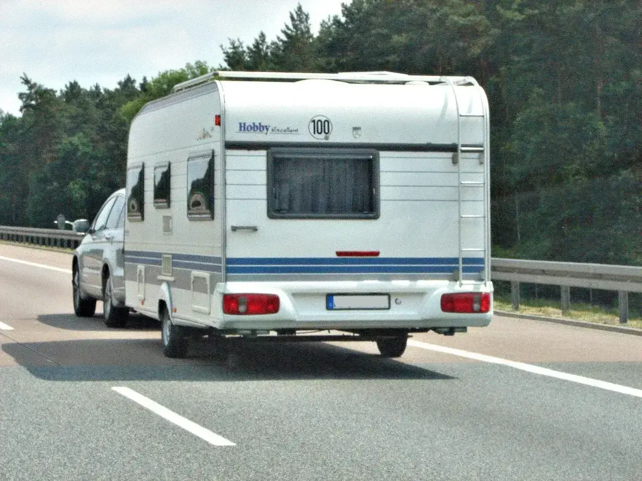 Biała przyczepa kempingowa Hobby z niebieskim pasem jedzie autostradą. Kierowca z prawem jazdy kategorii B+E wiezie rodzinę na wakacje.