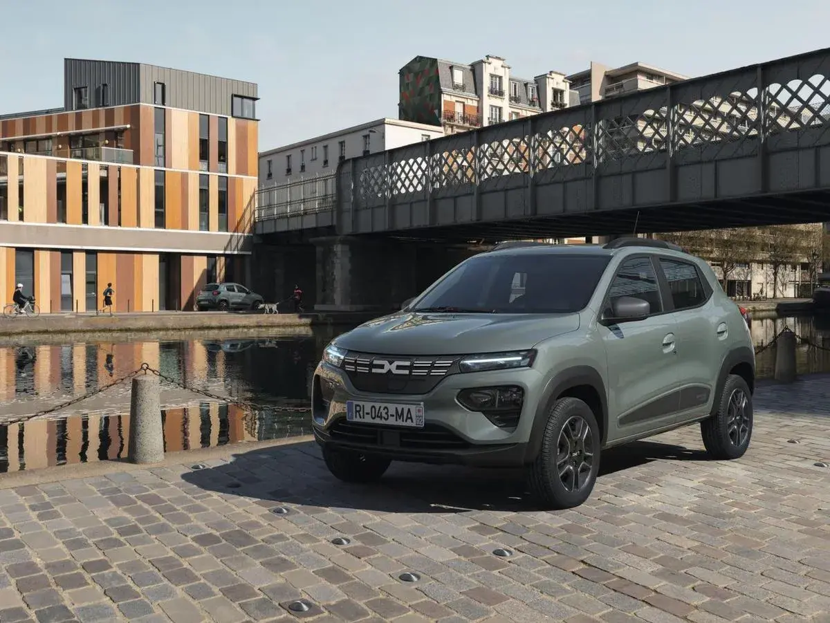 Dacia Spring cena: zielony elektryczny samochód miejski stoi na brukowanej ulicy nad kanałem, w tle nowoczesne budynki i most.