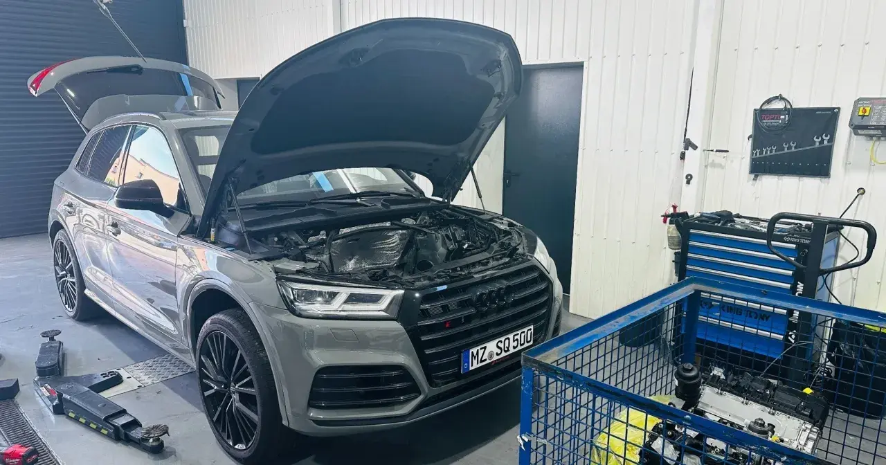 Szary Audi Q7 3.0 TDI z otwartą maską i bagażnikiem w warsztacie. Obok stoi wózek z narzędziami i silnik.