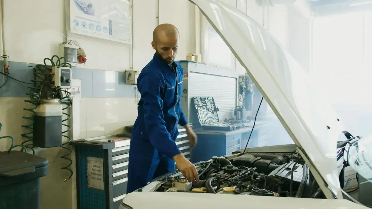 Mechanik w niebieskim kombinezonie sprawdza silnik samochodu, z którego wydobywa się dym. Obok stoi skrzynka z narzędziami i urządzenie przypominające parownik.