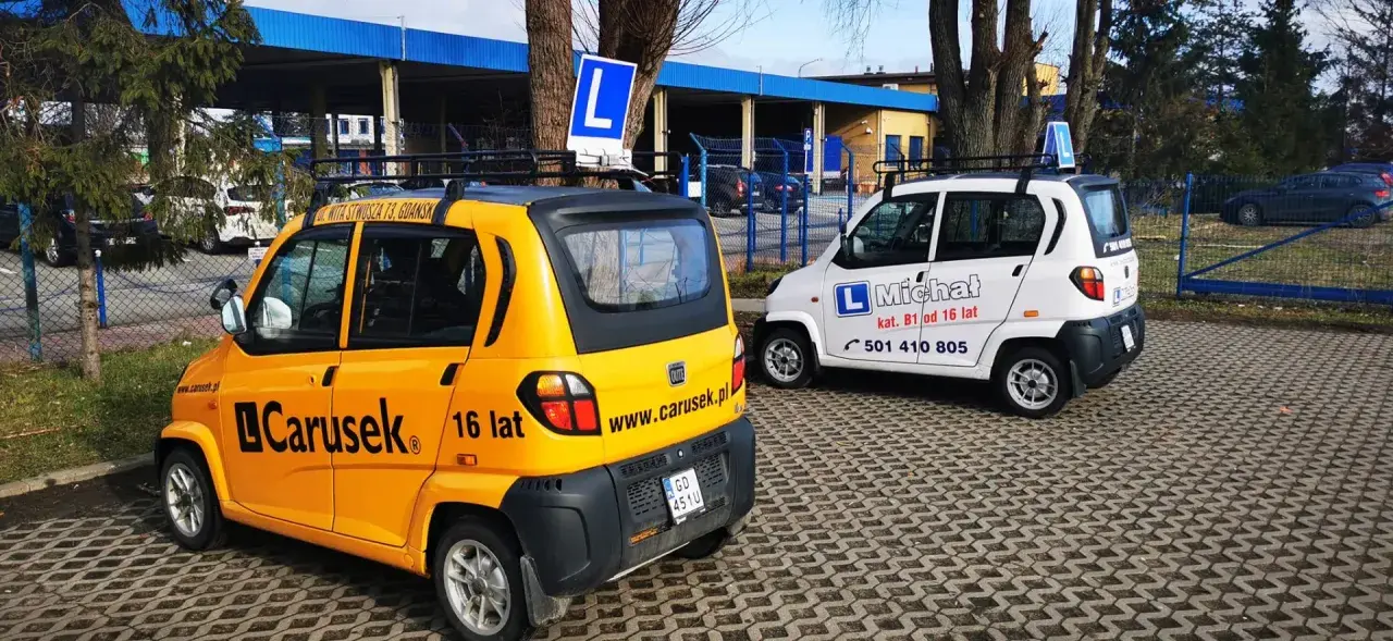 Dwa samochody szkoleniowe, żółty i biały, z literą "L" na dachu. Na żółtym aucie napis "Carusek 16 lat", sugerujący możliwość uzyskania prawo jazdy od 16 lat.