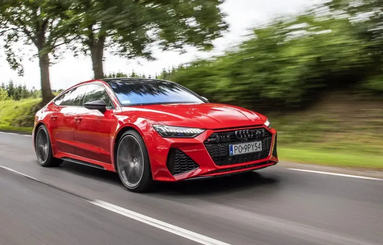 Czerwony Audi RS7 Sportback pędzi po drodze, otoczony zielenią drzew.