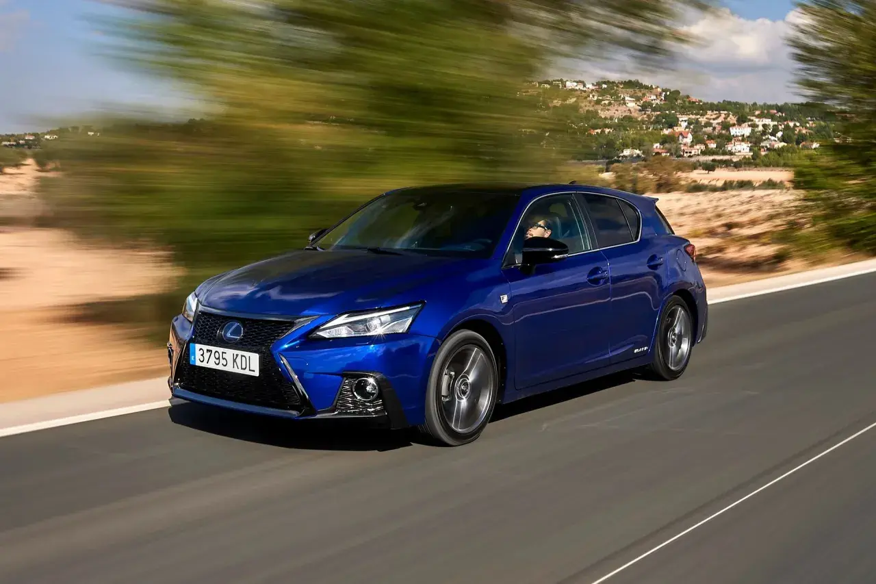 Błękitny Lexus CT 200h pędzi po krętej drodze, mijając zielone drzewa i domy na wzgórzu.